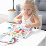 A young girl with blonde hair places colorful toys into Bumkins: Clear Travel Bag 3 Pack: Boots at a white table.