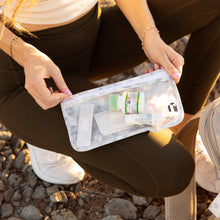 Load image into Gallery viewer, A person in athletic wear kneels on rocky ground, holding a Roamio by Bumkins Clear Travel Bag 3 Pack: Disco Check with first aid supplies.
