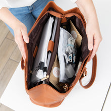 Load image into Gallery viewer, A person opens a brown purse on a white table, revealing notebooks and a Roamio by Bumkins Clear Travel Bag 3 Pack: Disco Check inside.
