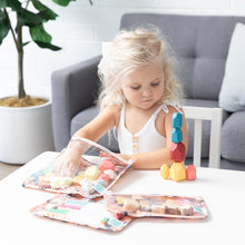 Load image into Gallery viewer, A blonde girl plays with blocks at a white table while reaching into Bumkins Clear Travel Bag 3 Pack: Yoga Pals filled with toys and art supplies.
