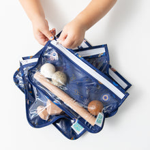 Load image into Gallery viewer, A child unzips a Bumkins Clear Travel Bag 3 Pack: Evil Eye with toys inside on a white table.
