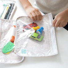 Load image into Gallery viewer, Close-up of a child's hands zipping a Bumkins Clear Travel Bag 3-Pack: Wander packed with toy cars and small items, resting on a white table.
