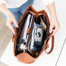 Load image into Gallery viewer, A person opens a brown purse on a white table, revealing notebooks and a Roamio by Bumkins Clear Travel Bag 3 Pack: Crushed Denim inside.
