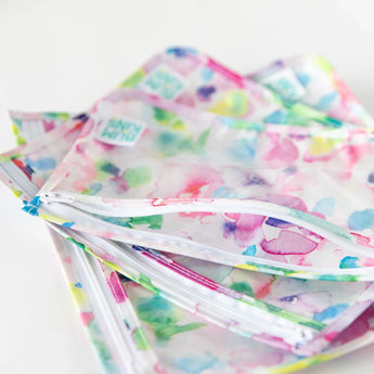 Close-up of Bumkins Clear Travel Bag 3-Pack: Watercolor stacked on a white background, with the top back unzipped, revealing the interior.