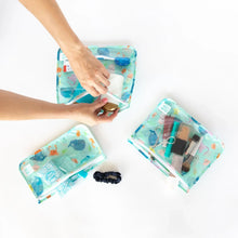 Load image into Gallery viewer, Hands packing TSA-approved items and makeup into Bumkins Clear Travel Bag 3-Pack: Ocean Life on a white background.
