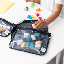 Load image into Gallery viewer, A child's hand reaches for blocks from a Bumkins Clear Travel Bag 3-Pack: Mickey Mouse Icon Black + White at a white table with art supplies.

