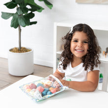 Load image into Gallery viewer, A curly-haired girl smiles at a white table, reaching into a Bumkins Clear Travel Bag 3 Pack: Aloha Stitch filled with toys.
