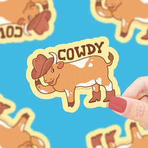 Sticker of a cowboy cow with COWDY on blue backdrop. Vinyl, premium quality by Turtle Soup. Hand with red nails holds it.