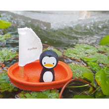 Load image into Gallery viewer, A toy penguin from PlanToys sails in a PlanWood orange boat with a white sail on a pond surrounded by lush leaves.
