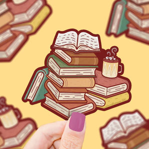 Vinyl Sticker, Cozy Book: books stacked messily with a cup of hot cocoa and marshmallows on top of two books; held over a yellow background.