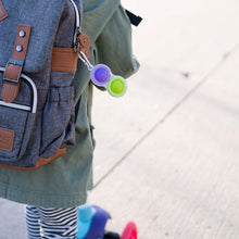 Load image into Gallery viewer, A child in a green jacket holds a backpack with a Fat Brain Toy Cos Simpl Dimpl. A colorful scooter is nearby on the sidewalk.
