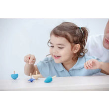 Load image into Gallery viewer, A young child smiles while playing with PlanToys Spinning Tops, enhancing fine motor skills, as an adult supervises.
