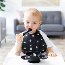 Load image into Gallery viewer, In a high chair, a toddler smiles in a Bumkins Silicone Bib: Mickey Mouse Faces, holding a spoon with a bowl in front.

