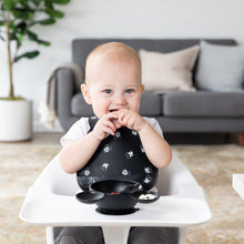 Load image into Gallery viewer, A baby in a high chair wears a Bumkins Silicone Bib: Mickey Mouse Faces, enjoying snacks from a black bowl. A grey sofa and plant sit behind.

