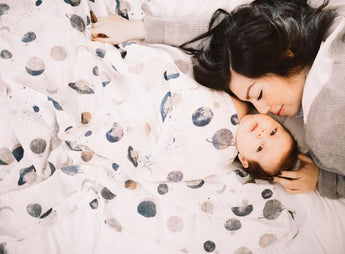 A woman and baby snuggle under Little Unicorns breathable Cotton Swaddle, Planetary blanket.